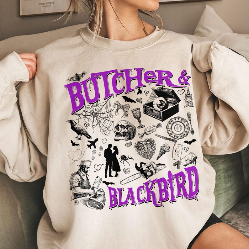 May include: Beige sweatshirt with a purple graphic that reads "Butcher & Blackbird." The design includes a spiderweb, skull, crow, and other spooky illustrations. A unique and stylish top for Halloween or everyday wear.