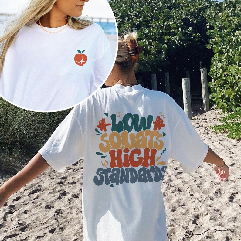 May include: White t-shirt with a red apple graphic on the front and the text "Low Squats High Standards" in colorful, stylized lettering on the back.