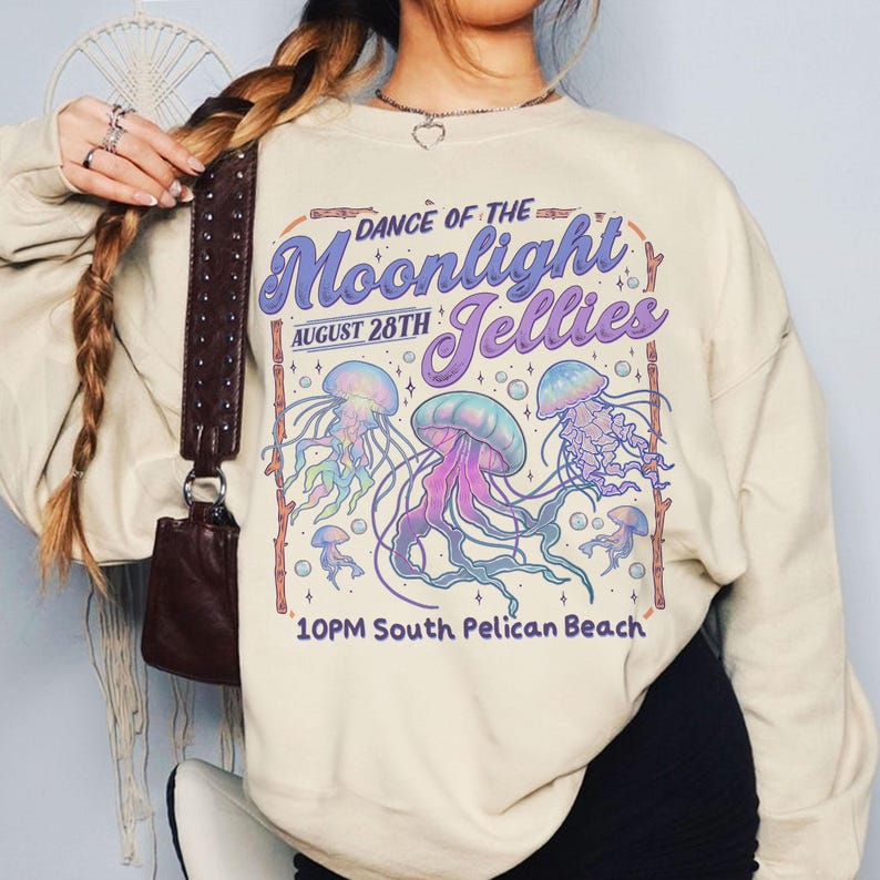 Puede incluir: Sudadera de color crema con un dise&ntilde;o gr&aacute;fico. El dise&ntilde;o presenta el texto "Dance of the Moonlight Jellies" y la fecha del 28 de agosto, junto con ilustraciones de medusas coloridas. La parte inferior del dise&ntilde;o dice "10PM South Pelican Beach."