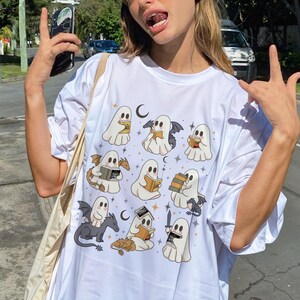 May include: White t-shirt with a playful design of cartoon ghosts reading books, with bats, dragons, and crescent moons. The ghosts are in various poses, some with wings, and holding books. A fun and unique graphic tee.