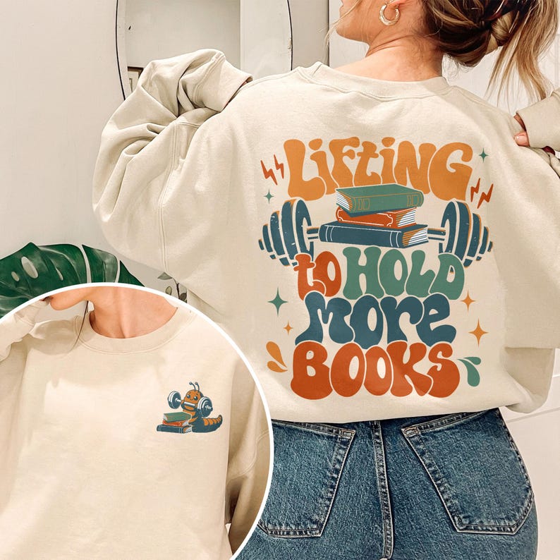 May include: A cream-colored sweatshirt with the text "Lifting to hold more books" in a retro font. The text is accompanied by an illustration of a barbell with a stack of books on it. The sweatshirt is being worn by a person with long brown hair and blue jeans.