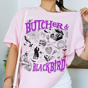 May include: A light pink t-shirt with the words "BUTCHER & BLACKBIRD" in purple, along with various black and white illustrations. The illustrations include bats, a spiderweb, a skull, and a chainsaw.