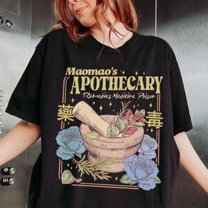 May include: Black t-shirt featuring a vintage apothecary design. The graphic includes a mortar and pestle with herbs, blue roses, and the text "Maomao's Apothecary Remedies Medicine Poison" in a retro style. The design has a botanical and medicinal theme.