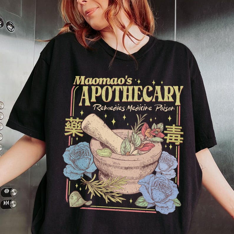 May include: Black t-shirt featuring a vintage apothecary design. The graphic includes a mortar and pestle with herbs, blue roses, and the text "Maomao's Apothecary Remedies Medicine Poison" in a retro style. The design has a botanical and medicinal theme.