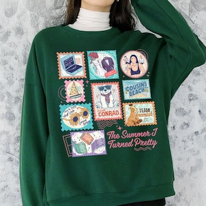 May include: Dark green sweatshirt with a collage of stamp-like graphics. The stamps feature illustrations of a laptop, a couple, a woman, a sailboat, a bear, and text that reads "Cousins Beach", "Team Conrad", and "Team Jeremiah". The text "The Summer I Turned Pretty" is also visible.