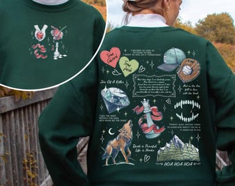 May include: A dark green crewneck sweatshirt featuring a small graphic on the front with an apple, a rose, a ribbon, and a chess piece. The back displays a large collage of illustrations including hearts, a baseball cap, a glove, a diamond, a wolf, and text.