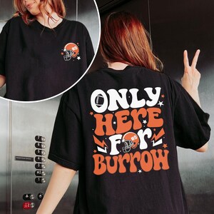 以下が含まれることがあります： 前面にフットボールのヘルメットのグラフィック、背面に「ONLY HERE FOR BURROW」の文字が入った黒いTシャツ。文字はレトロなフォントで、オレンジと白の色を使用しています。
