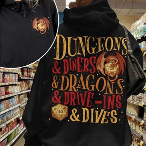 May include: Black hoodie with the text "DUNGEONS & DINERS & DRAGONS & DRIVE-INS & DIVES" in gold and red. A dragon and dice graphic is centered on the back. A smaller version of the graphic is on the front left chest.