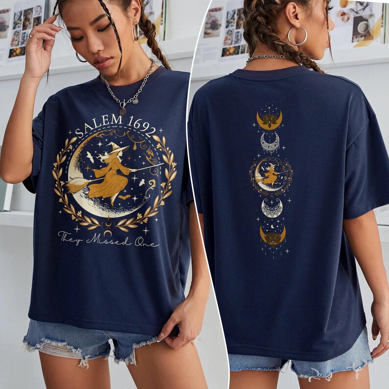 May include: A navy blue t-shirt featuring a graphic of a witch flying on a broomstick in front of a crescent moon. The text "Salem 1692" and "They Missed One" is printed on the shirt. The back of the shirt has a graphic of five moons with stars and a witch flying on a broomstick.