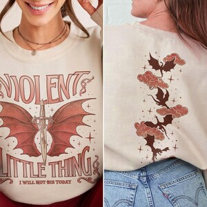 May include: A cream-colored sweatshirt with a graphic print of a red dragon with wings and a sword. The text "VIOLENT LITTLE THING I WILL NOT BE SHE TODAY" is printed below the dragon. The back of the sweatshirt has a graphic print of four red dragons flying in the clouds.