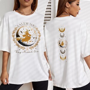 May include: White t-shirt with a gold graphic of a witch flying on a broomstick with the text "Salem 1692 They Missed One". The back of the shirt has a gold graphic of a crescent moon with stars.