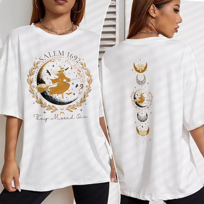 May include: White t-shirt with a gold graphic of a witch flying on a broomstick with the text "Salem 1692 They Missed One". The back of the shirt has a gold graphic of a crescent moon with stars.