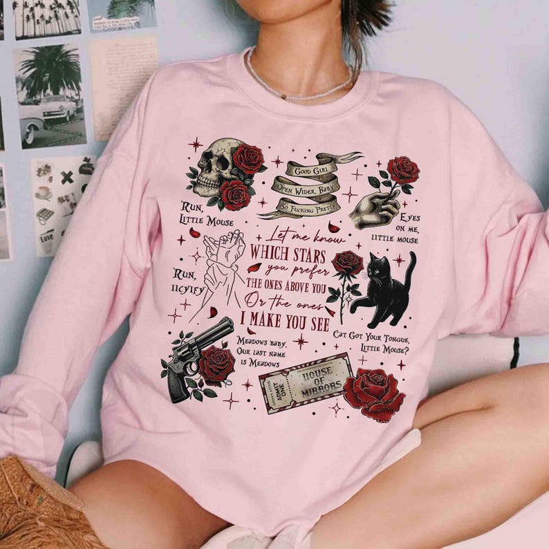 May include: Pink sweatshirt with a graphic print of a skull, roses, a cat, a gun, and text that says "Let me know which stars you prefer. The ones above you or the ones I make you see."