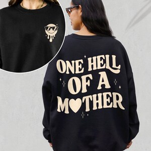 May include: Black sweatshirt with a white graphic on the back that says "One Hell of a Mother" with a heart in the word "Mother". The front of the sweatshirt has a small white graphic of a smiling face with sunglasses.
