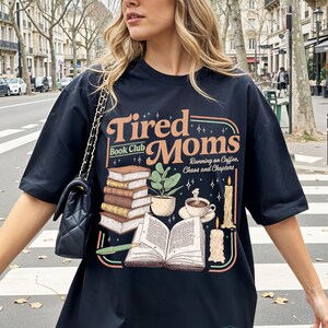 Puede incluir: Camiseta negra con el texto "Tired Moms Book Club" en un dise&ntilde;o retro. El gr&aacute;fico incluye pilas de libros, una planta en maceta, una taza de caf&eacute; y velas. Tambi&eacute;n incluye la frase "Running on Coffee, Chaos and Chapters".