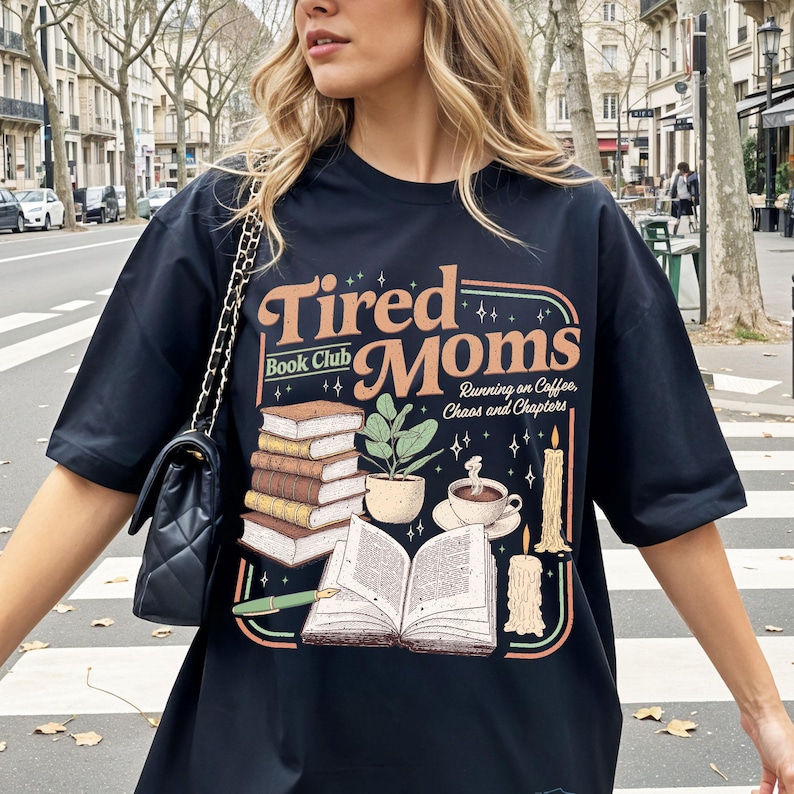Puede incluir: Camiseta negra con el texto "Tired Moms Book Club" en un dise&ntilde;o retro. El gr&aacute;fico incluye pilas de libros, una planta en maceta, una taza de caf&eacute; y velas. Tambi&eacute;n incluye la frase "Running on Coffee, Chaos and Chapters".