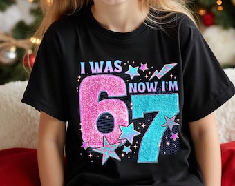 May include: A black short-sleeved t-shirt featuring a graphic print with the text I WAS NOW I'M 67. The design includes a large pink glittery number 6 and a large blue glittery number 7, surrounded by various blue and pink star shapes.