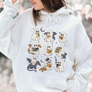 May include: White hoodie with a repeating pattern of cartoon ghosts reading books, with bats, dragons, and moons. The ghosts are in various poses, some holding books, others with wings or swords. The design includes stars and a crescent moon.