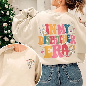 May include: Cream-colored sweatshirt with the text "IN MY DISPATCHER ERA" in colorful bubble letters. The design includes a walkie-talkie, headphones, and an SOS icon. A smaller version of the design is on the front, with the name "Kylie".
