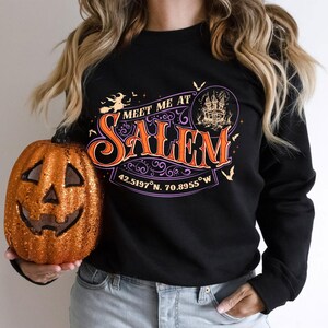 May include: A black long-sleeve sweatshirt featuring a graphic print with the text "MEET ME AT SALEM" in orange and purple. The design includes a witch on a broom, a haunted house, bats, and the coordinates 42.5197°N, 70.8955°W in white text.