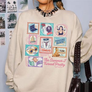 May include: Cream-colored sweatshirt featuring a colorful stamp design with various illustrations and text. The stamps include a laptop, wedding attire, a sailboat, a bear, and the words "Cousins Beach" and "Team Conrad". The text "The Summer I Turned Pretty" is also present.