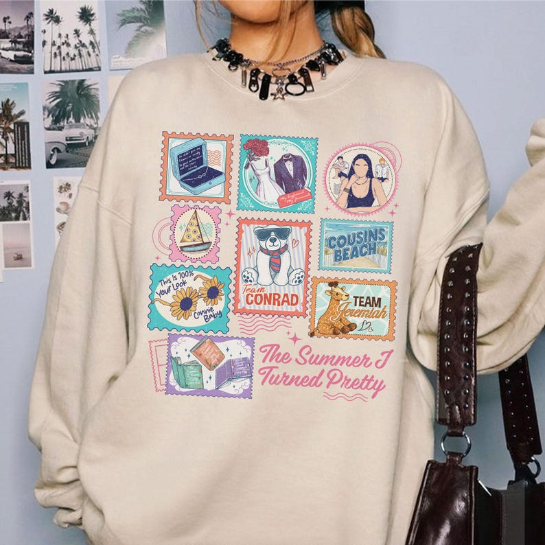 May include: Cream-colored sweatshirt featuring a colorful stamp design with various illustrations and text. The stamps include a laptop, wedding attire, a sailboat, a bear, and the words "Cousins Beach" and "Team Conrad". The text "The Summer I Turned Pretty" is also present.