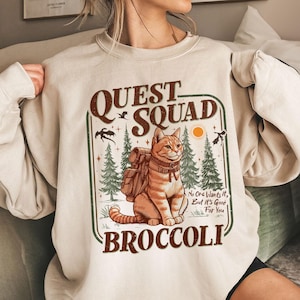 May include: A beige sweatshirt with a graphic print of a cat wearing a backpack and the text "Quest Squad Broccoli". The text below the cat reads "No one wants it, but it's good for you."