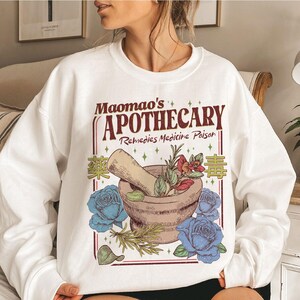 May include: White sweatshirt featuring a graphic design with the text "Maomao's Apothecary Remedies Medicine Poison". The design includes a mortar and pestle, blue roses, and botanical elements. The sweatshirt is a casual style.