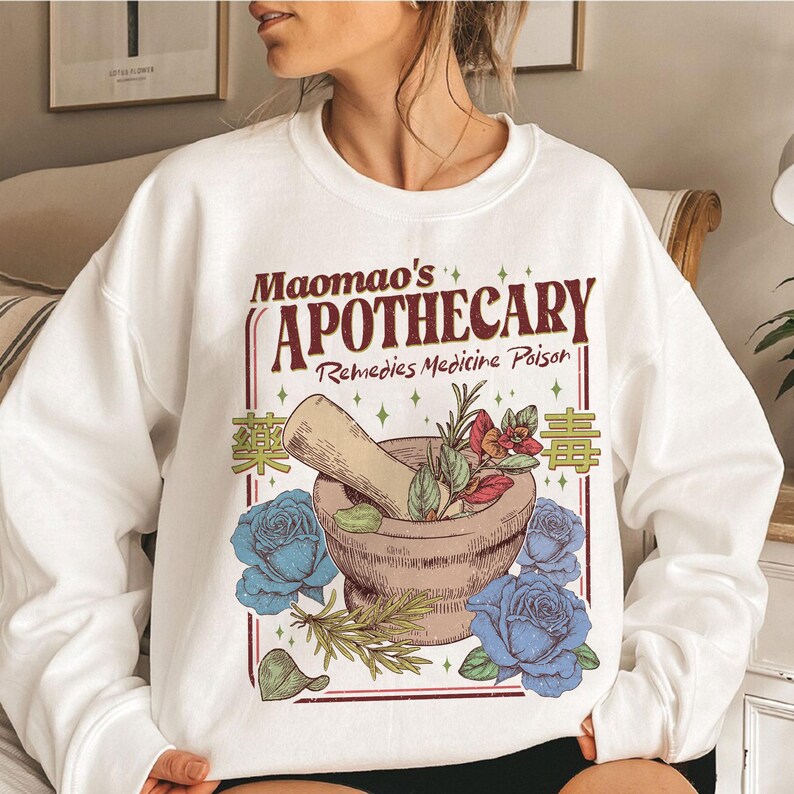 May include: White sweatshirt featuring a graphic design with the text "Maomao's Apothecary Remedies Medicine Poison". The design includes a mortar and pestle, blue roses, and botanical elements. The sweatshirt is a casual style.