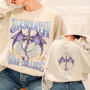 May include: Cream-colored sweatshirt featuring a purple dragon with outstretched wings, the text "BASGIATH" above and "WAR COLLEGE" below. The back of the sweatshirt has a similar dragon design. The design is in shades of purple, blue, and gold.