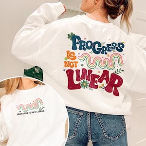 May include: White sweatshirt with the text "Progress is not Linear" in a colorful, whimsical design. The design includes a rainbow, flowers, and stars. The sweatshirt is a crew neck style.