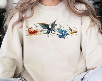 May include: A cream-colored crewneck sweatshirt featuring a graphic print of Santa Claus in a red sleigh being pulled by three flying dragons in shades of green, blue, and yellow, surrounded by small green snowflakes and sparkling star accents.