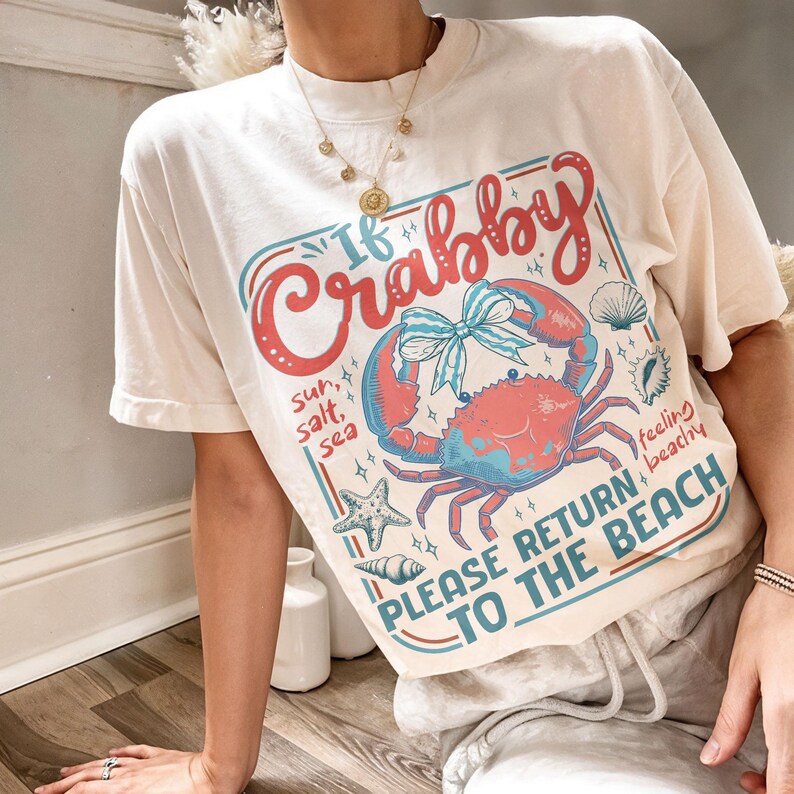 以下が含まれることがあります： カニ、貝殻、ヒトデのグラフィックが描かれたクリーム色のTシャツ。「If Crabby, Please Return to the Beach」と書かれています。カニは赤と青で、青いリボンが付いています。