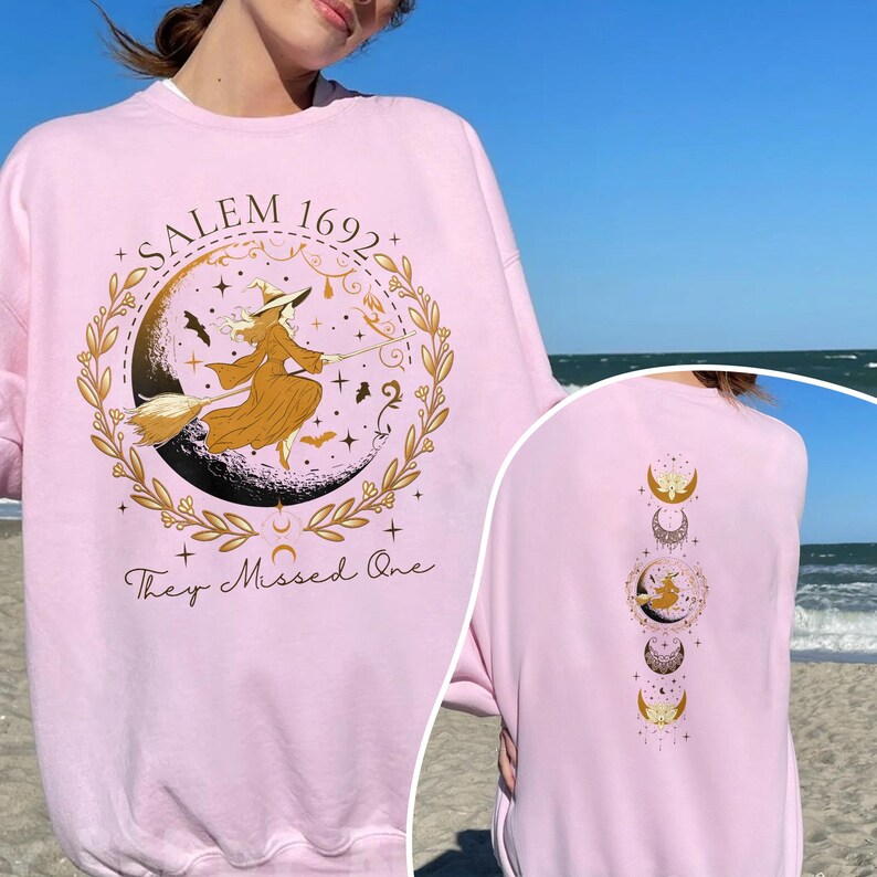 May include: A pink sweatshirt with a gold graphic of a witch flying on a broomstick with the text "Salem 1692 They Missed One".