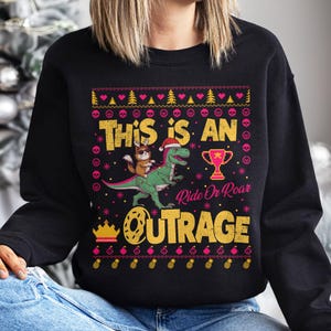 May include: Black sweatshirt with a colorful graphic design. The design features a dinosaur ridden by a cat wearing a Santa hat, a trophy, and the words "THIS IS AN OUTRAGE". The design also includes hearts, trees, and skulls.