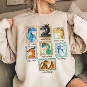 May include: Beige sweatshirt featuring a colorful stamp design with various dragon illustrations. Each stamp showcases a different dragon type, including NightWings, SeaWings, and SandWings, with corresponding text labels. The design is centered on the front of the sweatshirt.