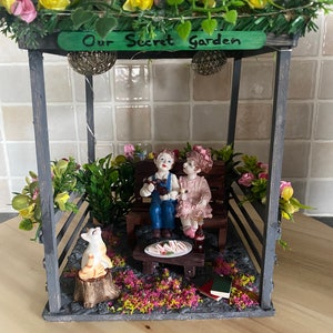 Può includere: Una scena di giardino in miniatura con un gazebo grigio, con una panchina con due statuette, un gatto e un tavolo con cibo. Il gazebo è adornato con fiori e il testo "Our Secret Garden".