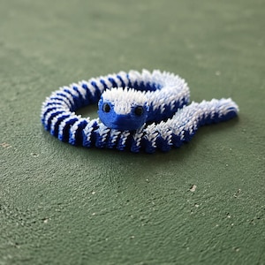 Op de afbeelding: Een 3D-geprint, beweegbaar slangenfiguur met een felblauw lichaam en een witte, stekelige textuur langs de bovenkant. De slang heeft twee kleine zwarte, ronde ogen en ligt in een cirkelvormige positie op een plat oppervlak.