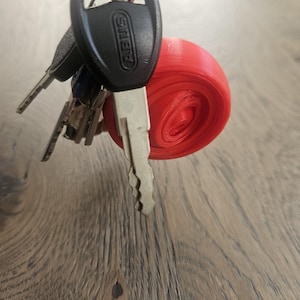 Op de afbeelding: Een sleutelbos bevestigd aan een zilveren sleutelring, met een rood, cilindrisch object. De sleutels omvatten een zwarte sleutel met het merk "ABUS" en andere sleutels van verschillende maten en vormen. Het rode object lijkt een 3D-geprint accessoire te zijn.