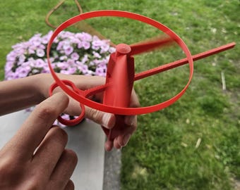 Peut inclure: Un jouet volant en plastique rouge vif doté d'un cadre circulaire et d'un mécanisme d'hélice central. L'appareil comprend une poignée avec un anneau pour le doigt permettant une utilisation manuelle, conçue pour lancer l'hélice en l'air.