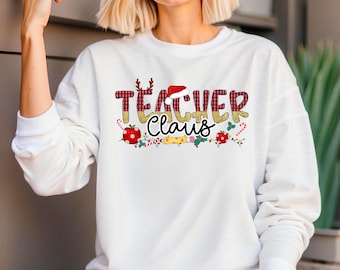 Könnte beinhalten: Weißes Langarm-Sweatshirt mit festlichem "Teacher Claus" Design. Der Text hat ein Karomuster, einen Weihnachtsmannhut und Rentiergeweihe. Dekorative Elemente sind Zuckerstangen, Äpfel und Stechpalmen.