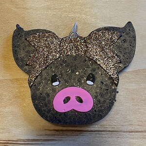 May include: A black pig ornament with a gold glitter bow. The pig has a pink snout and white eyes.