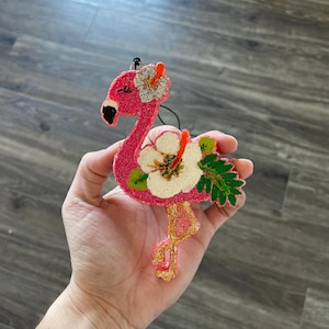 May include: Pink flamingo Christmas ornament with glitter and a white hibiscus flower. The flamingo has gold glitter accents on its legs and beak.