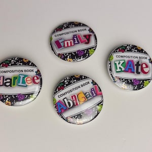 May include: Four round composition book-themed badges with names: Harlee, Emily, Kate, and Abigail. Each badge has a black and white design with colourful text and a white border. The badges are on a white surface.