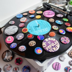 May include: A collection of colorful pinback buttons arranged on a black vinyl record, displayed on a vintage record player. The buttons feature various designs, including smiley faces, animal prints, and text. A small potted plant is visible in the background.