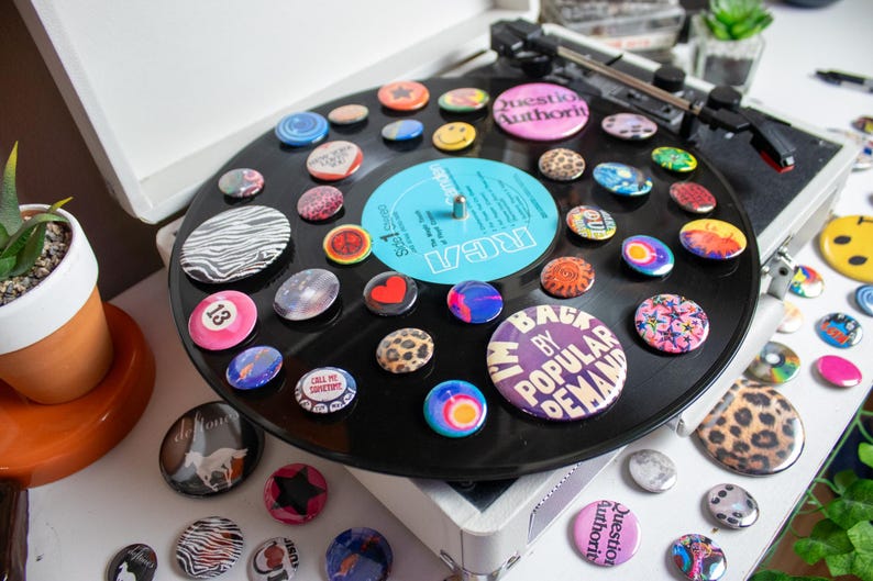 May include: A collection of colorful pinback buttons arranged on a black vinyl record, displayed on a vintage record player. The buttons feature various designs, including smiley faces, animal prints, and text. A small potted plant is visible in the background.