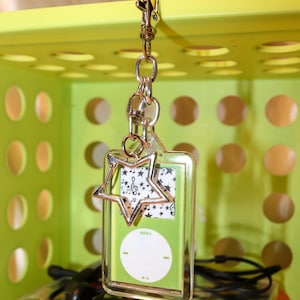 May include: A clear acrylic keychain shaped like a rectangular music player with a lime green face, featuring a white circular control pad with the text MENU, play, pause, and skip icons, accented by a silver star charm and a metal lobster clasp.