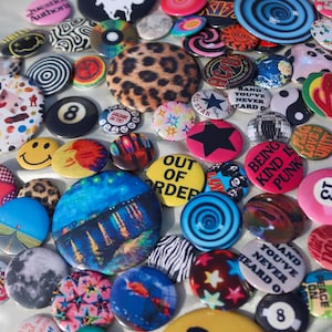 May include: A collection of colorful button pins with various designs. The pins feature images of a leopard print, a smiley face, a black star, and text that reads "OUT OF ORDER" and "BEING KIND IS PUNK".
