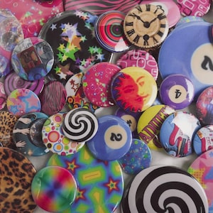 May include: A collection of colorful, round buttons with various designs. Some buttons feature swirls, leopard print, and celestial patterns. Others display a clock face, a billiard ball, and the word "POSER".