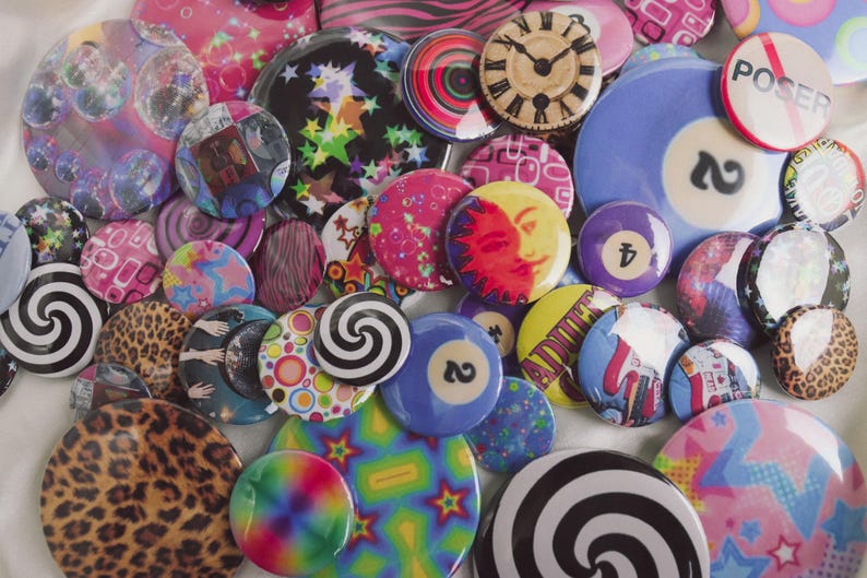 May include: A collection of colorful, round buttons with various designs. Some buttons feature swirls, leopard print, and celestial patterns. Others display a clock face, a billiard ball, and the word "POSER".